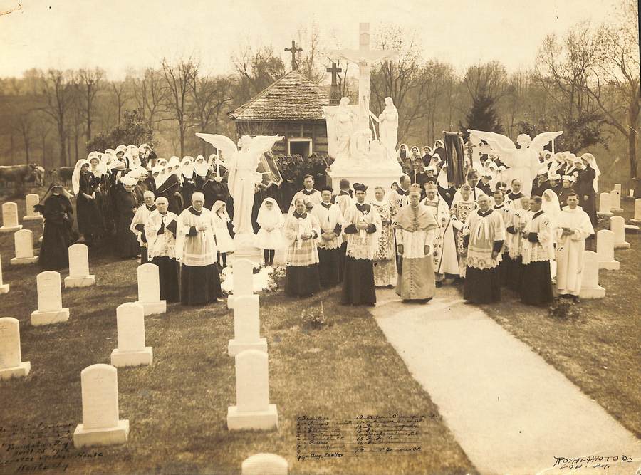 Archival photo in sepia tones of a group photo of religious in the Loretto Motherhouse cemetery. Multiple rows of sisters in habit stand at the back, with clergy in full cassocks and accoutrements in the foreground.