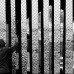 A black and white photo of somebody in a hoodie, shorts and sneakers back to the camera, putting their hands on the border wall between the U.S. and Mexico.