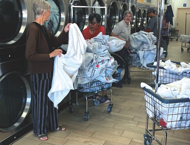 Three women are shown at a laundromat folding several baskets worth of white blankets.