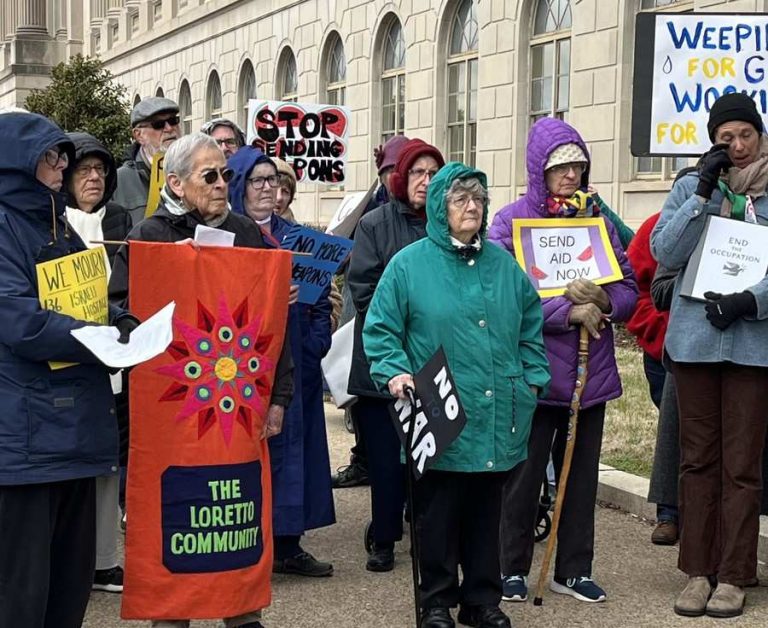 Women in winter coats stand outside a large building, holding signs in protest of the war in Gaza.