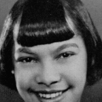 A black and white headshot picture of a young woman with straight black hair with bangs. She is smiling brightly in front of a plain background.