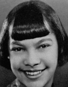 A black and white headshot picture of a young woman with straight black hair with bangs. She is smiling brightly in front of a plain background.