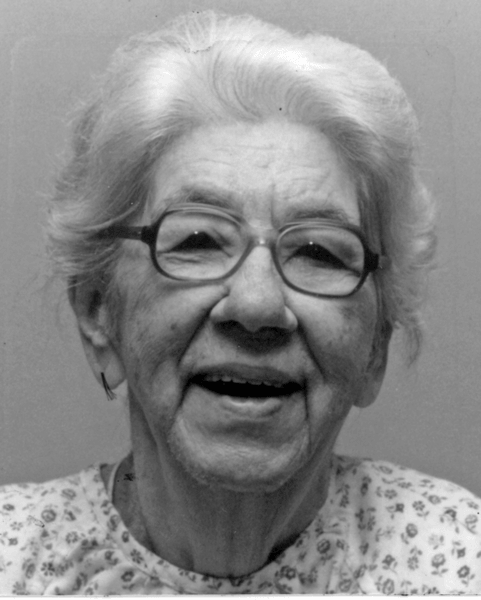 A black and white headshot picture of a woman with short, light-colored hair and oval-framed glasses smiling brightly wearing a white and floral patterned shirt in front of a plain background.