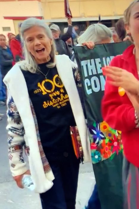 A woman smiling brightly wearing a warm jacket outdoors with her hand on a large poster sign amongst a crowd.