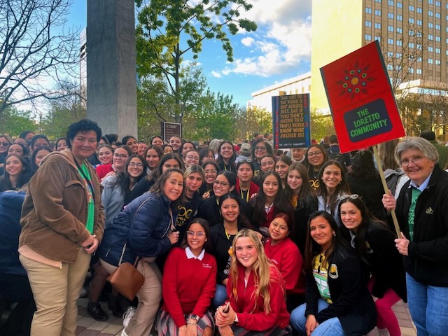 A crowd of mostly high school girls and a few adults take a photo at a protest. There are a couple signs being held by community members.