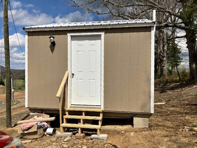 A small 16 x 16 light brown shed with a white door and three wood stairs leading up to it sits on concrete bricks.