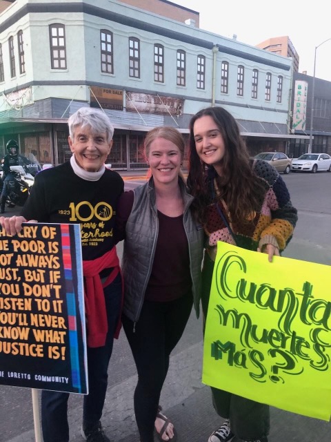 Three women, each of different generations smile together in a group photo at a protest, two of them are holding protest signs.