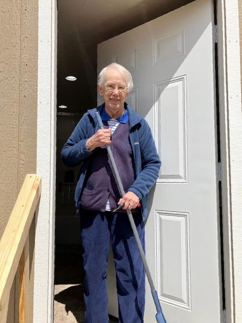 An older woman with white hair stands in the doorway of a shed holding a broom looking at the camera.