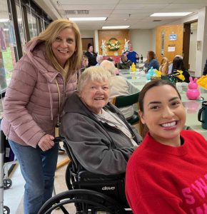 Three women smiling for a photo. One woman stands behind an older woman in a wheelchair and another woman sits at the communal table.