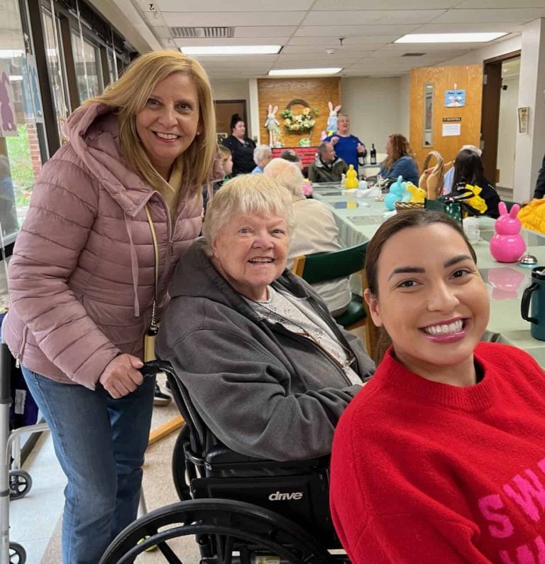 Three women smiling for a photo. One woman stands behind an older woman in a wheelchair and another woman sits at the communal table.