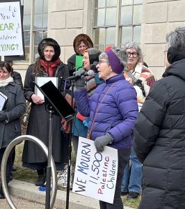 Pat Geier CoL, dressed for cold weather, speaks at a microphone to a group of people gathered outside a building.