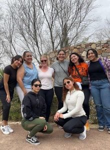 Eight women smile for a group photo on a casual hike on a cloudy day with six women standing in the back and two women squatting in the front wearing sunglasses.