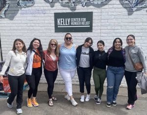Eight woman stand in a group photo smiling in casual clothing in front of a sign that says "Kelly Center for Hunger Relief"