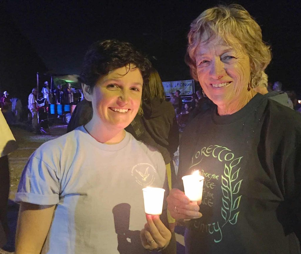 Two women stand outside in the night, holding votive candles.