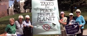 Six women outdoors on a sunny day wearing sunglasses and hats while standing next to and holding a large poster stating: Taxes for Peace People Planet.
