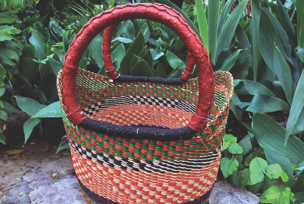 A brightly colored red, blue, green and white woven basket is placed outside with green plants around it.