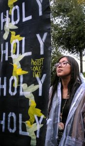 A young indigenous girl with big glasses, long dark hair and a necklace with a shell on it stands powerfully holding a sign that says "Holy Holy Holy Holy Protect Oak Flat."