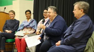 Five women sitting together in chairs on a small stage, wearing dress clothes. One woman is speaking into a microphone while the others listen.