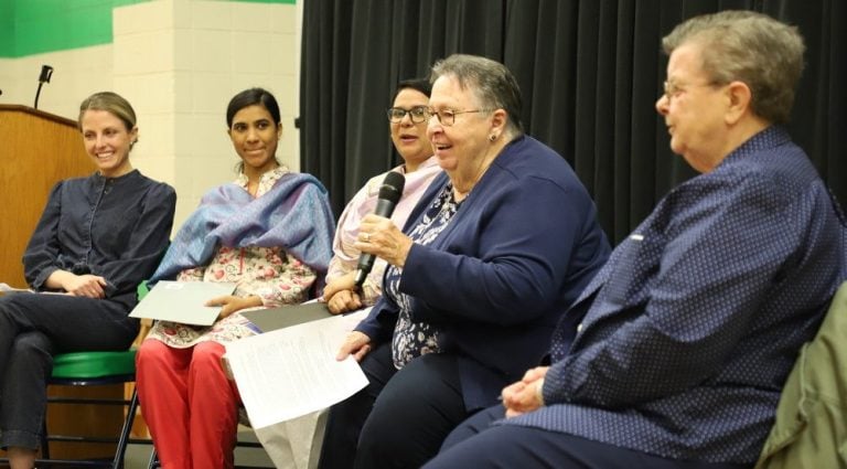 Five women sitting together in chairs on a small stage, wearing dress clothes. One woman is speaking into a microphone while the others listen.