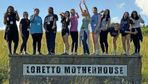 12 students pose silly while standing on a large stone sign that says: Loretto Motherhouse outdoors in front of a field.