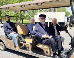 Four people smile together while riding on a golf cart outside on a sunny day.