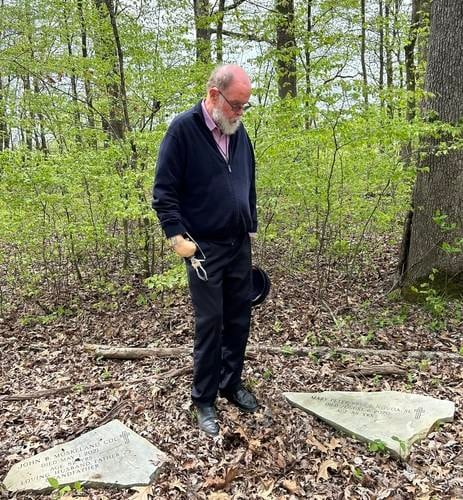 A man standing looks down at two headstones in a dense green forest.