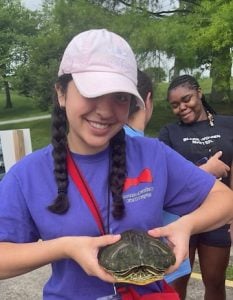 A woman in a pink hat in two braids and a purple shirt, smiling holding a turtle.