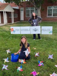 An elementary student sits on the ground amongst pinwheels. A teacher stands behind her and a sign that reads "PINWHEELS FOR PEACE." The o in "for" is a peace sign.