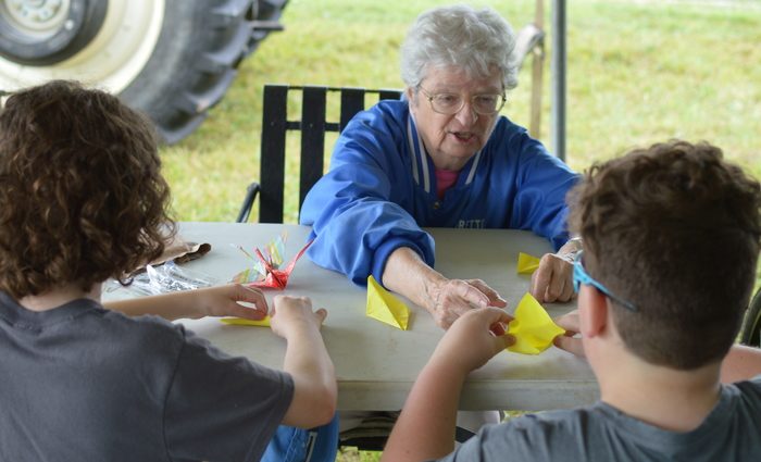 A woman demonstrates for two teens how to fold a paper crane at table at an outdoor event.