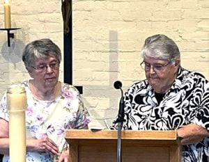 One woman watches as another one signs a paper on a wooden podium with a microphone on a church stage.