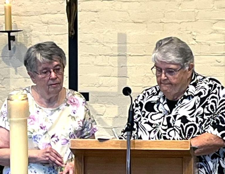 One woman watches as another one signs a paper on a wooden podium with a microphone on a church stage.