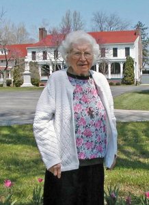 A woman with short white hair wearing a floral sweater and white cardigan smiling for a picture outdoors on a sunny day.