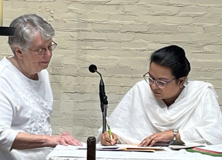 One woman supervises as another woman signs a paper at a podium with a microphone indoors.