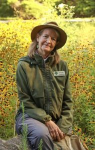 A woman smiling while kneeling in a field of sunflowers on a bright day.