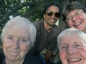 Four friends smile together for a selfie shaded by a large tree on a sunny day.