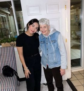 A young latina woman dressed in all black with black hair smiles standing next to a smiling older white woman with short grey hair, black pants and a jean jacket.