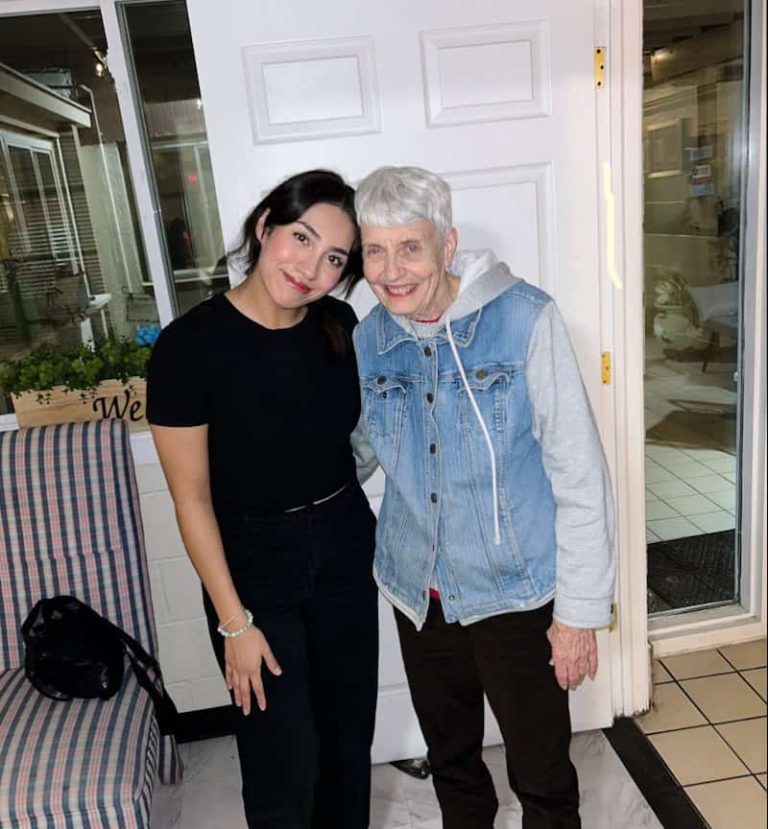 A young latina woman dressed in all black with black hair smiles standing next to a smiling older white woman with short grey hair, black pants and a jean jacket.