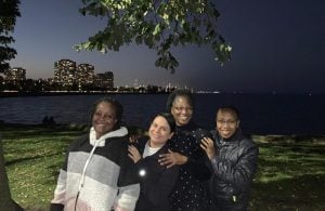 Four warmly dressed women of color pose for a night photo on the edge of a lake. Illuminated city buildings can be seen in the background, on the other side of the lake.