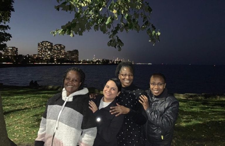 Four warmly dressed women of color pose for a night photo on the edge of a lake. Illuminated city buildings can be seen in the background, on the other side of the lake.