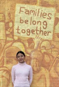 A latina woman with her dark hair pulled back, wearing a pink turtleneck, smiles softly in front of a mural that shows people marching and a sign that says "Families belong together."