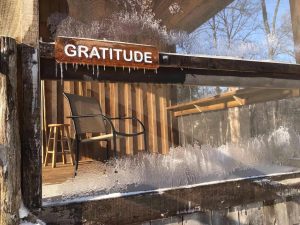 A sign that says "Gratitude" hanging on the outside of an icy wooden cabin window. Inside the window is a view of a chair on an indoor porch.