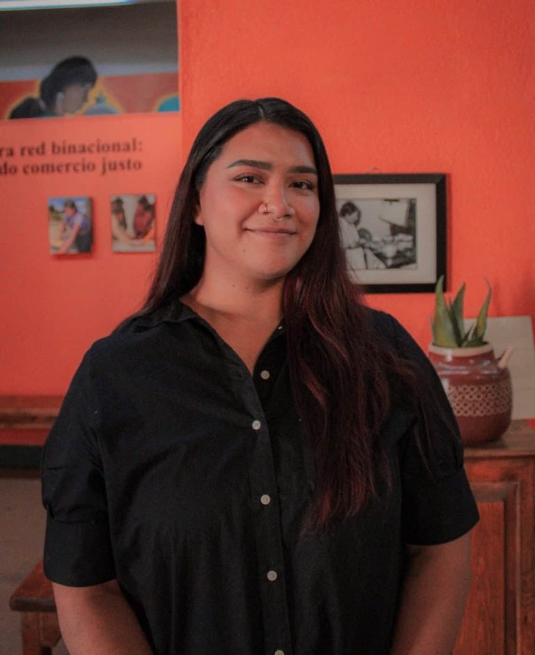 A latina woman smiles softly in front of a bright red wall with photos. She is wearing a black shirt and has long brown hair as well as a nose ring and septum ring.