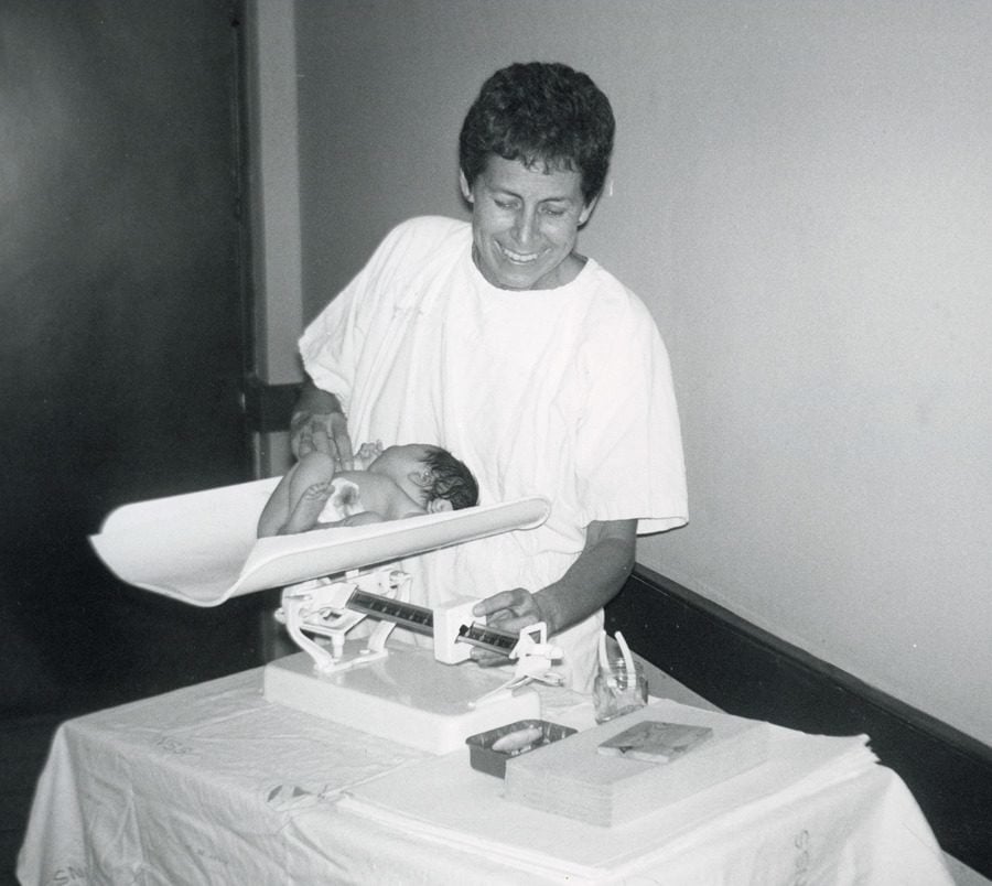 A woman in a white shirt weighs a small infant on a scale.