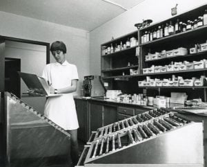 Black and white photo of a woman in a white uniform making notes on a medical chart in a dispensary.