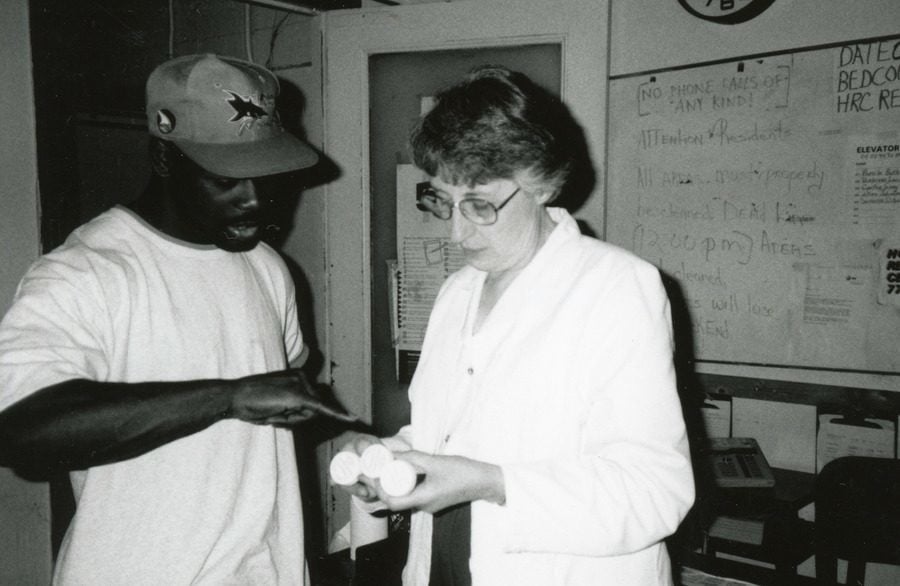 A White woman in a lab coat holds bottles of medication while listening to a Black man point something out on the bottle.