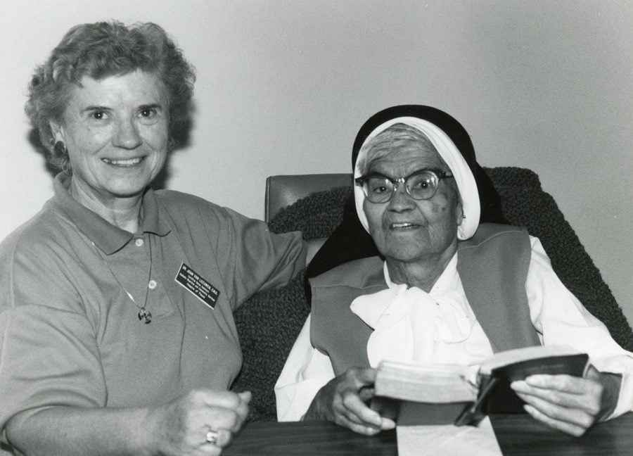 Black and white photo of a woman with a name tag kneeling next to a elderly nun seated in a comfortable chair.