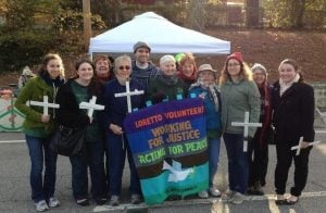 A group of mostly women and one man post for a photo holding crosses and a colorful banner that reads "Loretto Volunteers: Working for Justice, Acting for Peace."
