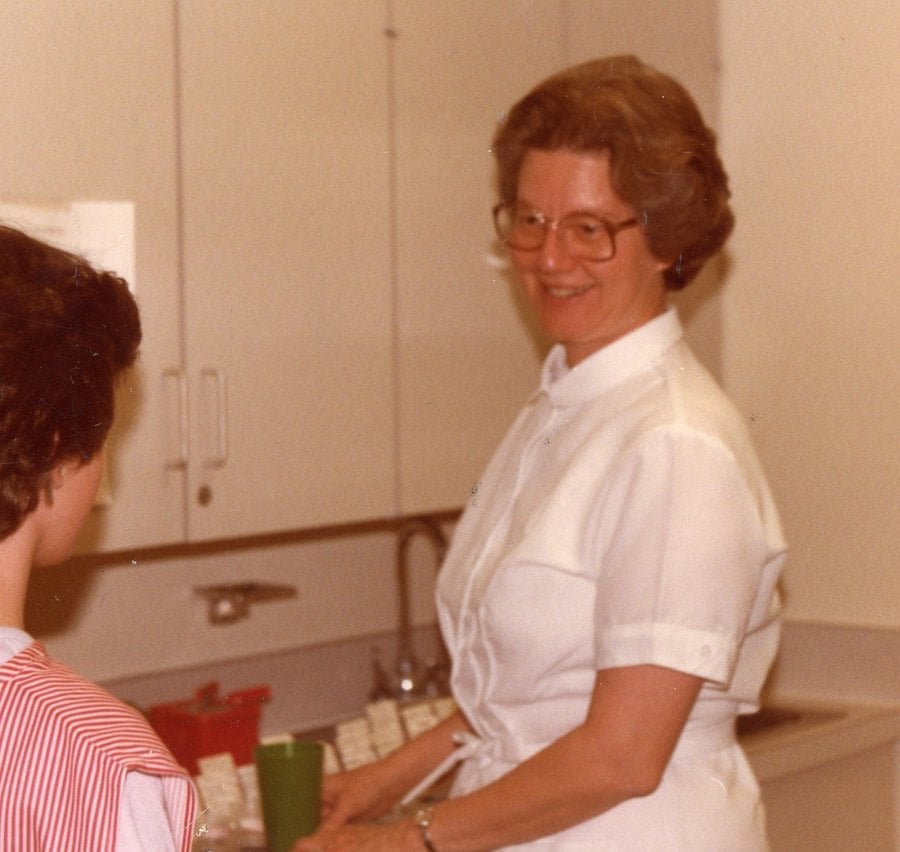 Color archival photo of a woman in a nurse's white uniform smiling at a candy striper.