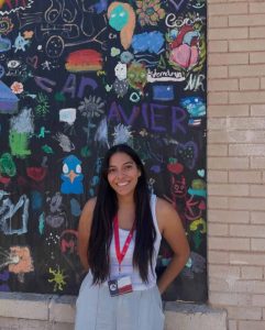 A hispanic woman with long waist length brown hair wearing light colored clothing stands smiling in front of a wall with several small pieces of colorful art all over.