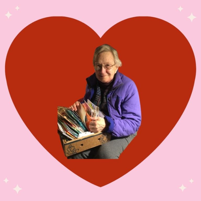 A red heart background surrounds Sister Roberta Hudlow as she sits with a box of letters.